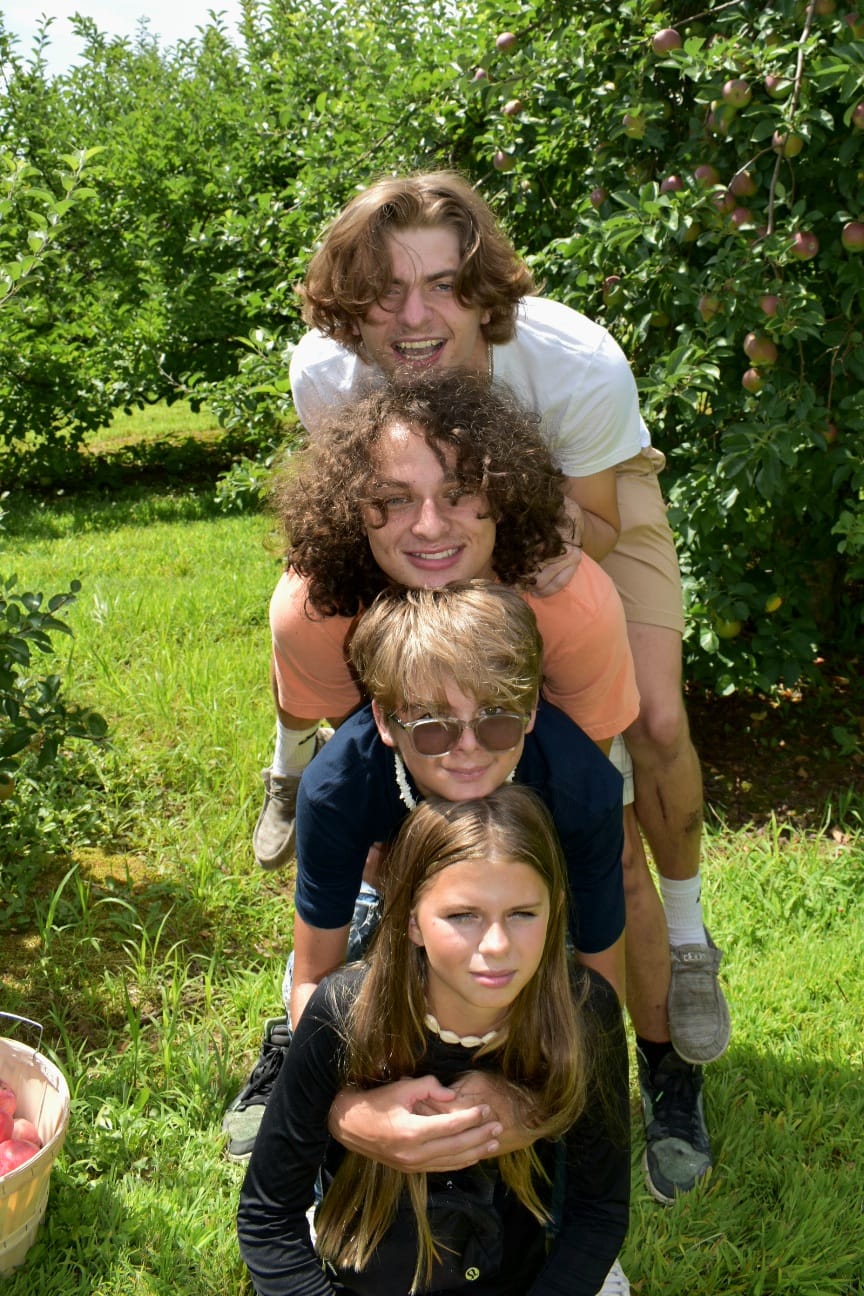 Four young people stacked in playful totem pole formation in apple orchard on sunny day, grinning and smiling