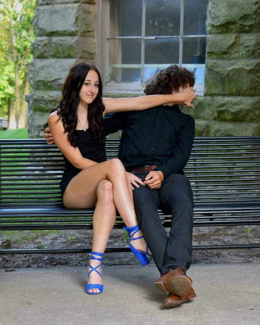 Two young people on park bench, woman in black dress playfully pushing companion's face while smirking
