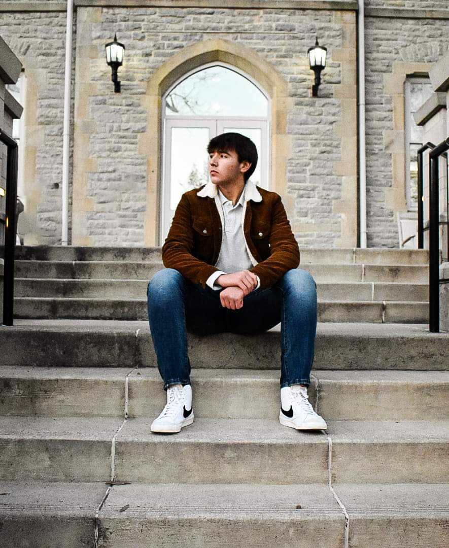Young man sits contemplatively on stone steps in front of gothic stone building wearing brown sherpa jacket