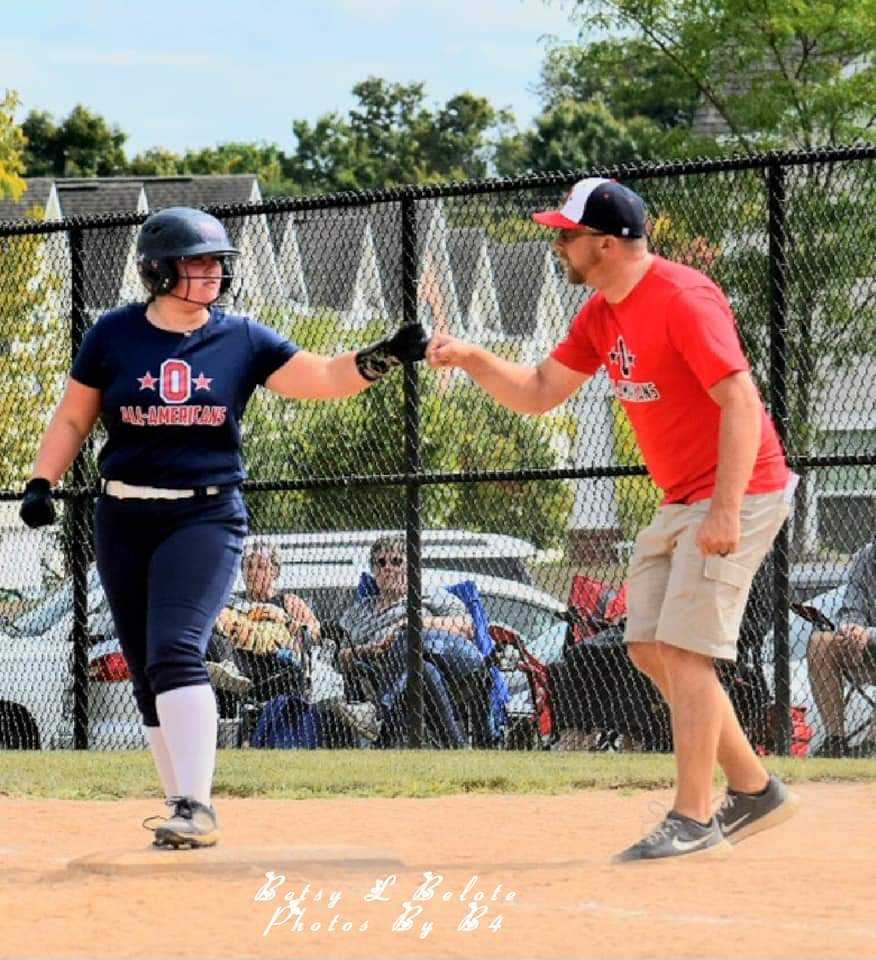 Youth softball player in navy blue All-Americans uniform fist-bumps her base coach near third base during game